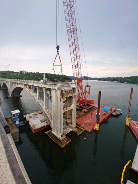 Large crane supporting bridge construction over a river with nearby barge and scenic forest backdrop