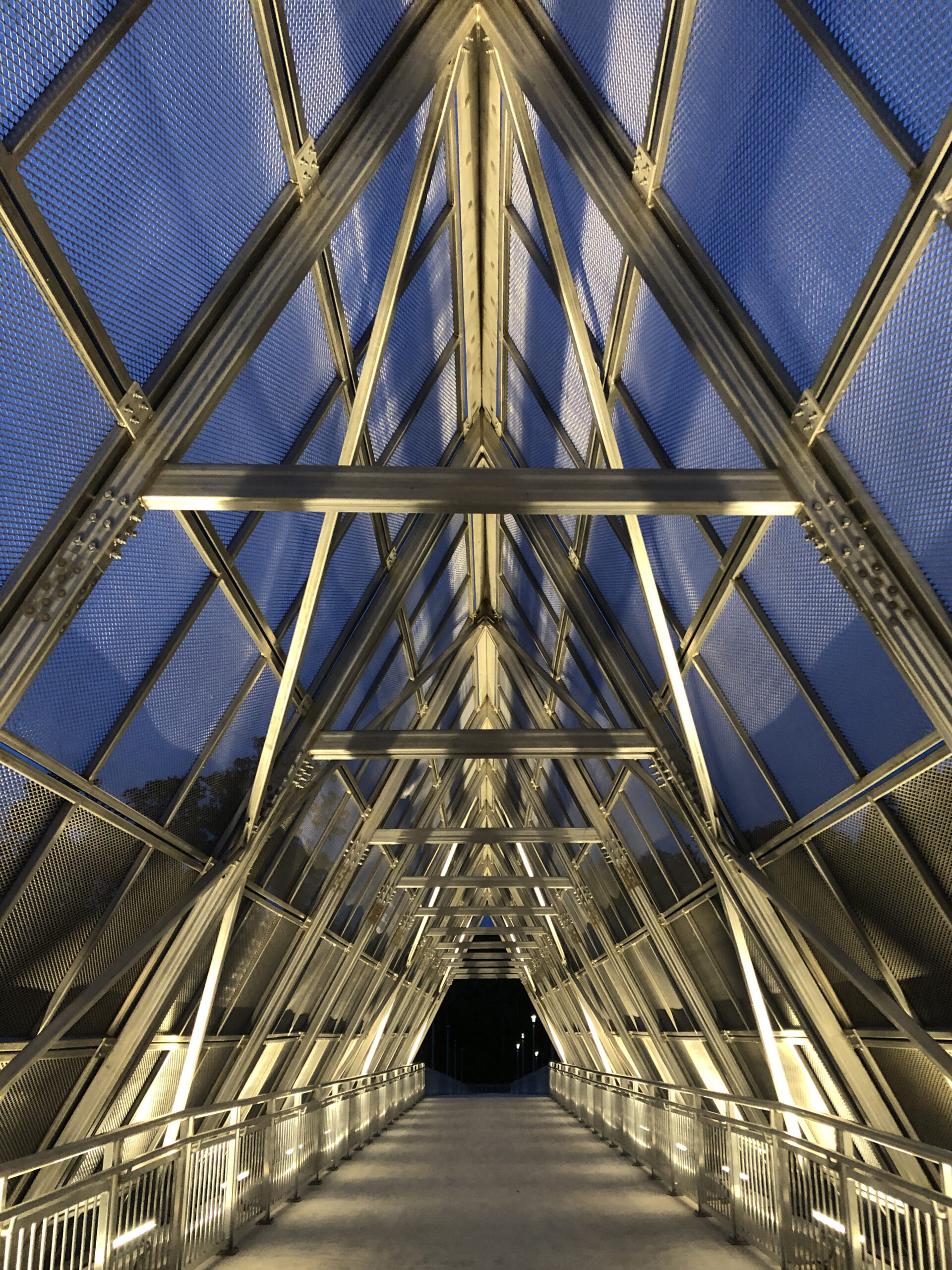 Metallic pedestrian bridge with illuminated triangular framework at dusk, creating a tunnel effect