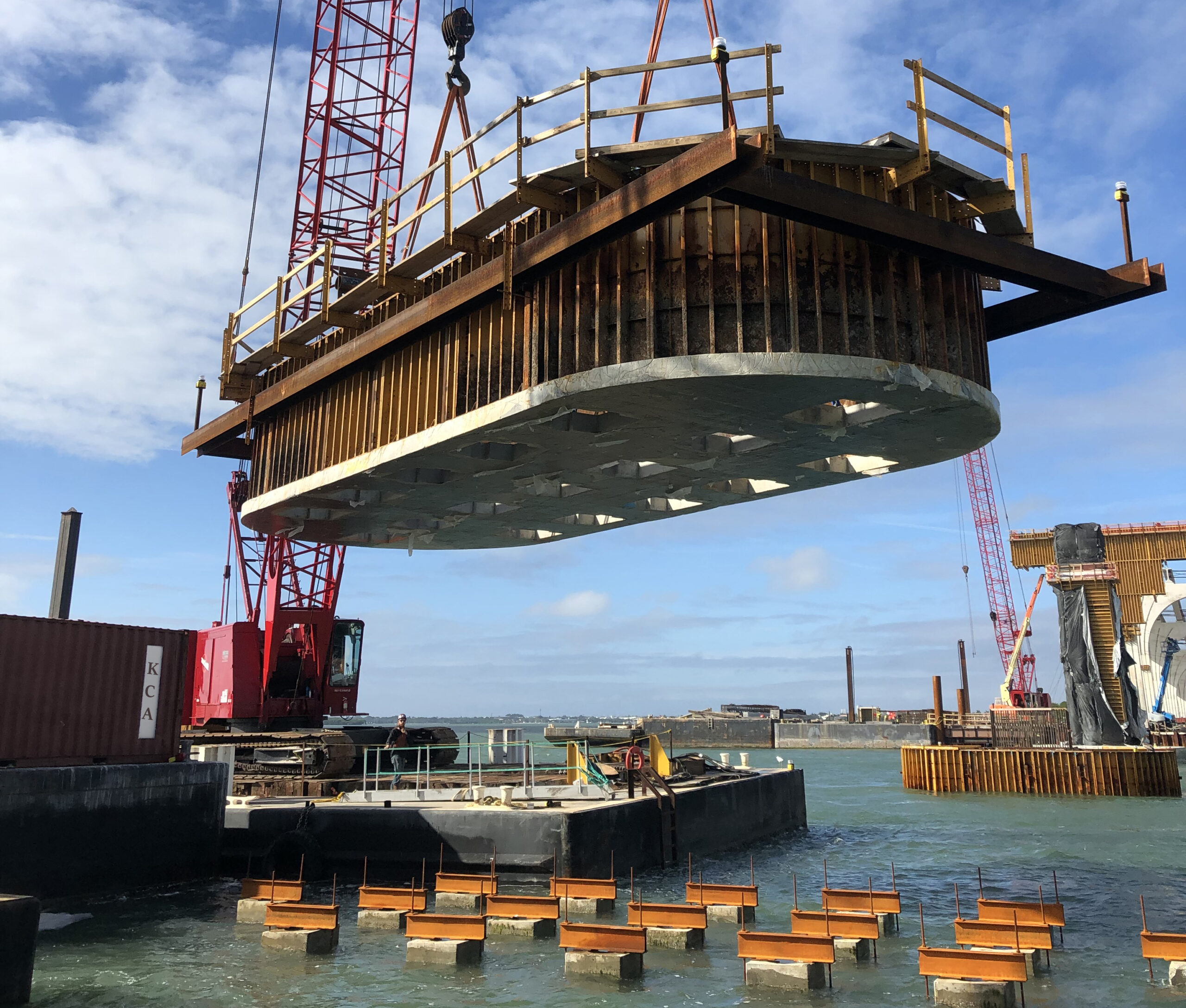 Large structure being lowered by crane onto water-based supports, with clear sky background