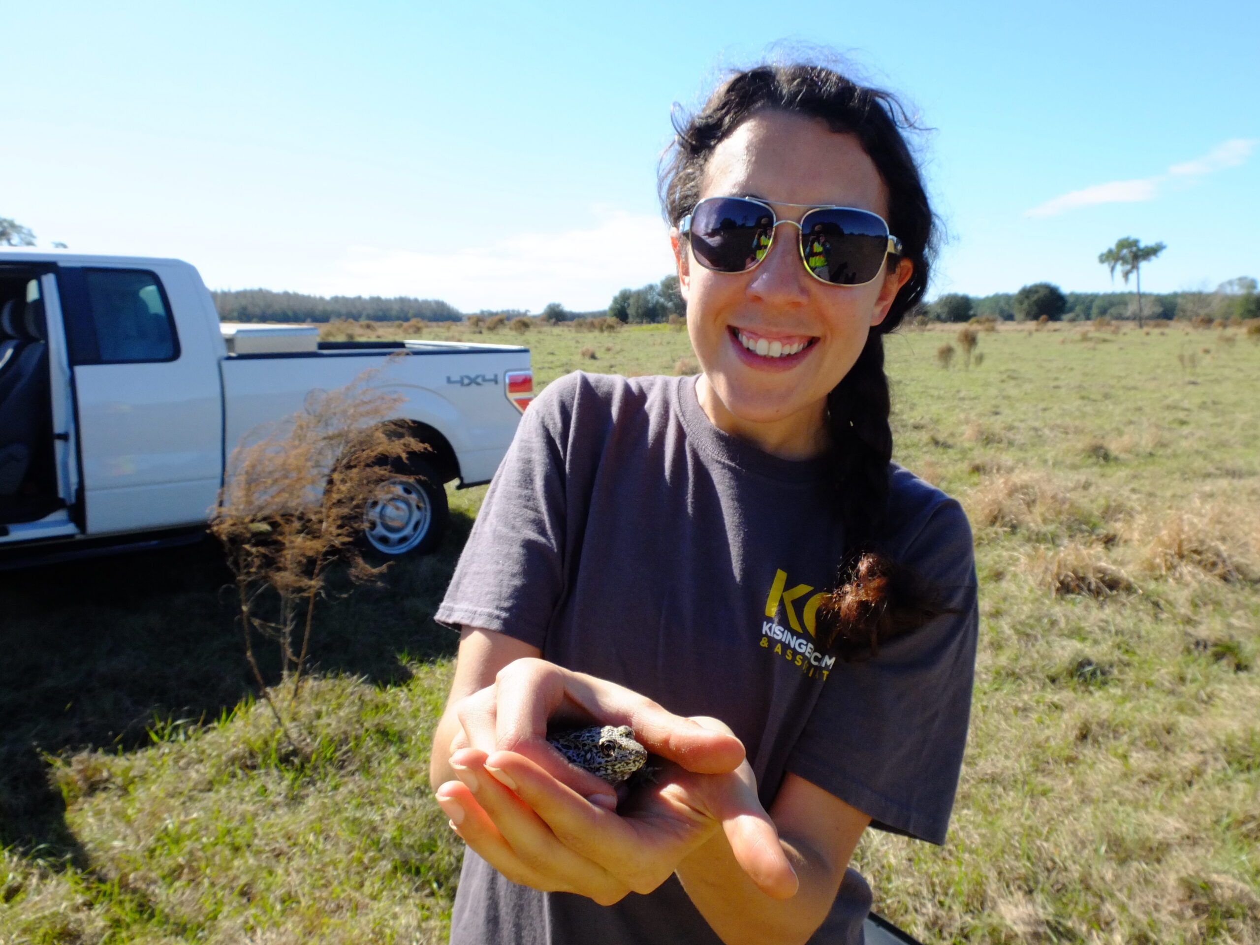 Smiling woman holding a small frog in a grassy field, truck in background