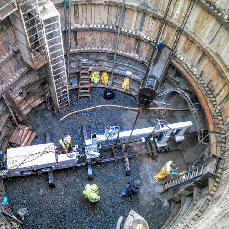Construction workers in a large cylindrical pit with machinery and ladders