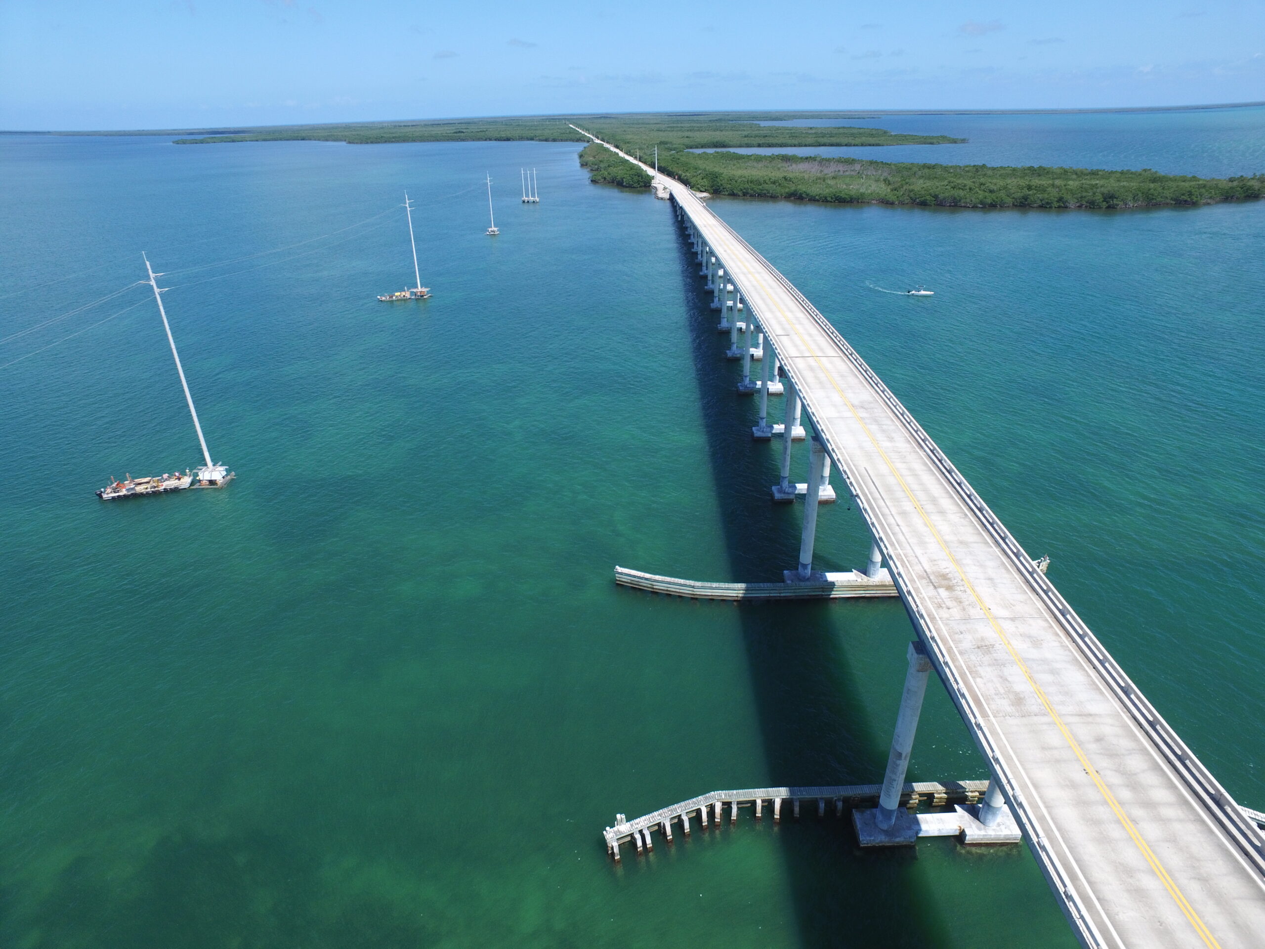 Aerial view of a long bridge over blue-green water, connecting two land areas with scattered sailboats nearby