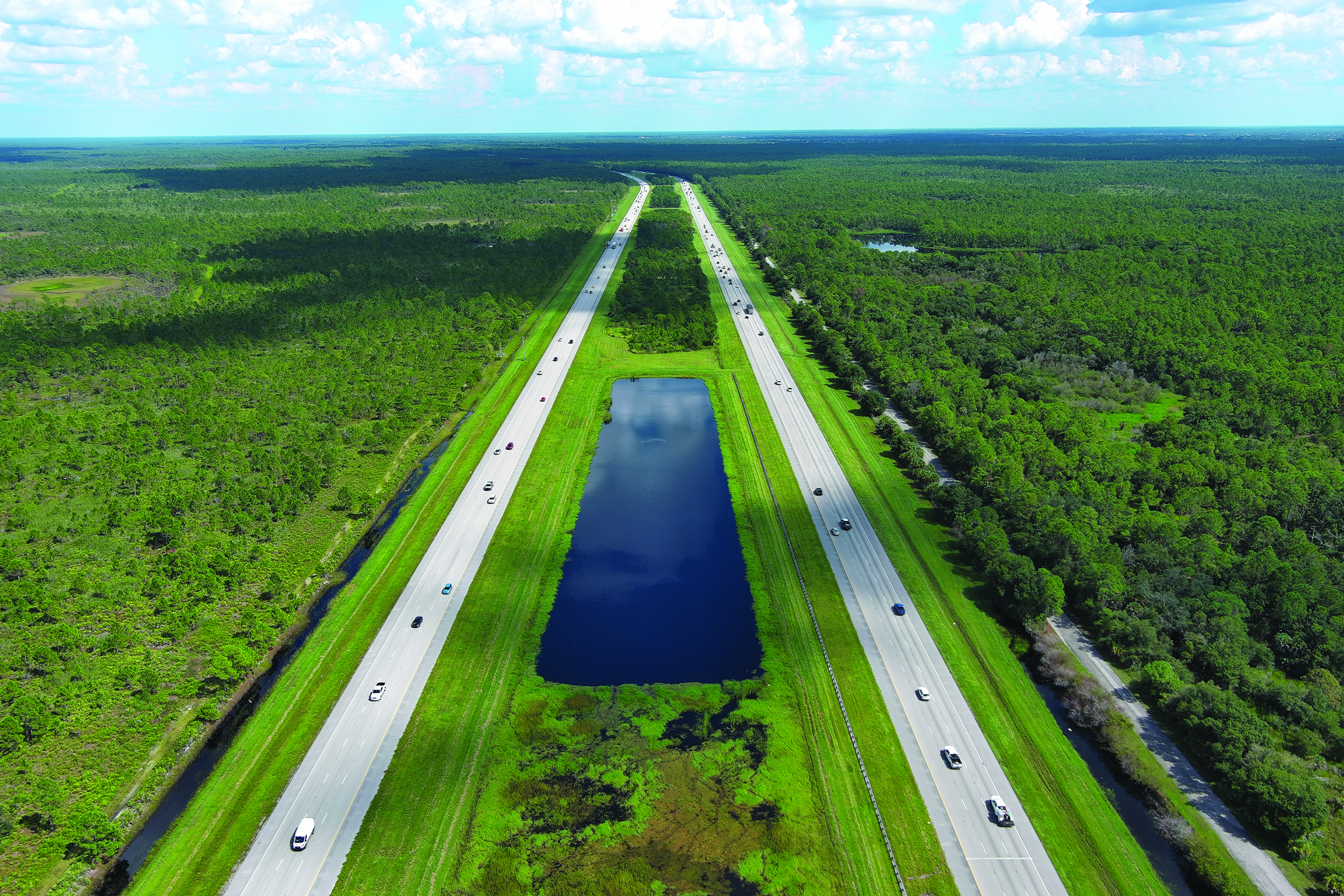 Aerial view of a divided highway with a water canal and dense green forest surrounding it