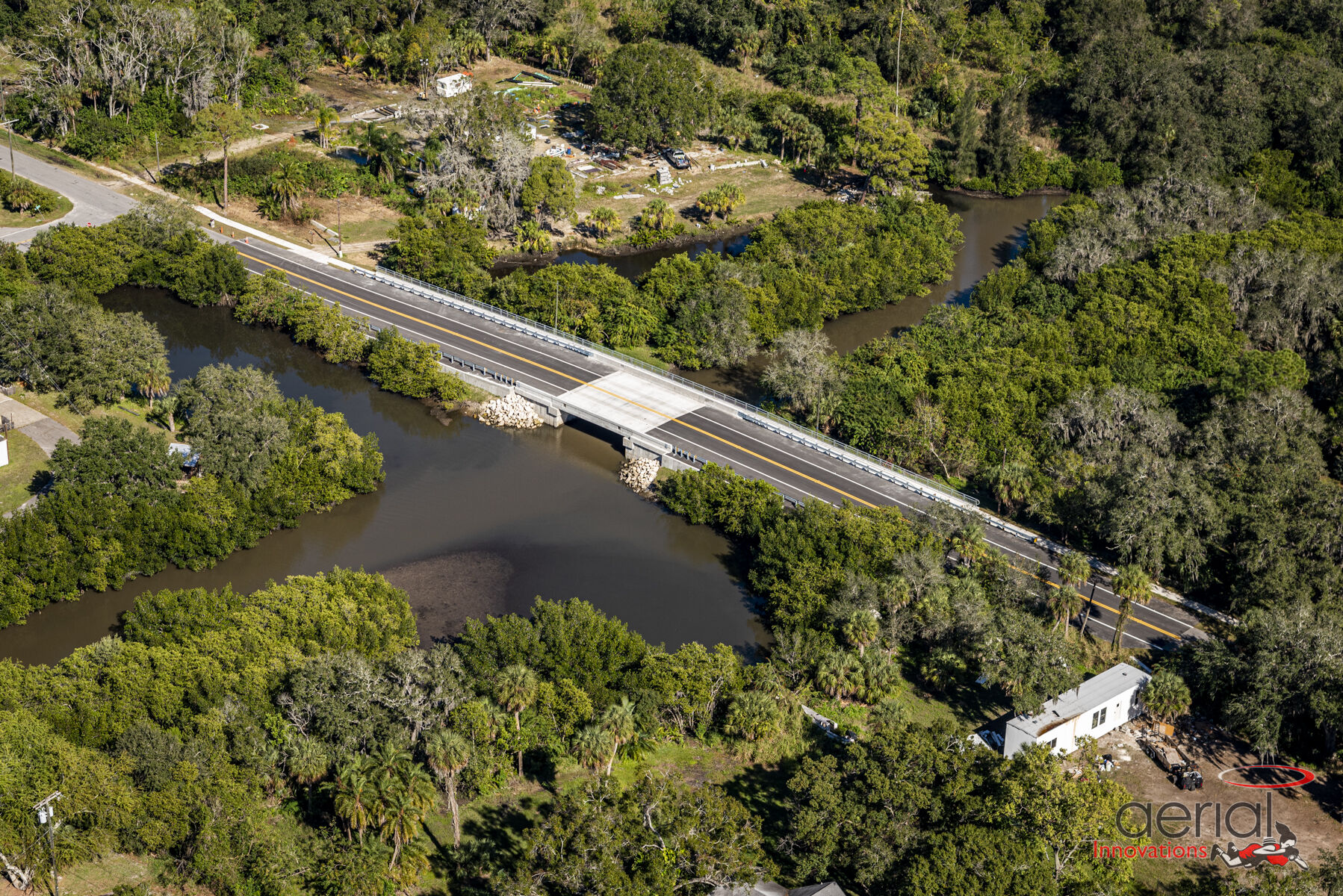 Aerial view of a bridge crossing a narrow river amidst dense green foliage, with a small white building nearby