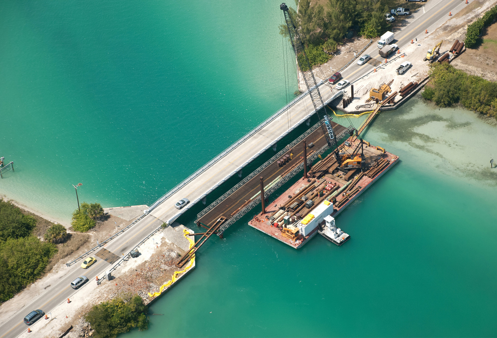 Aerial view of bridge construction over turquoise water, vehicles passing by