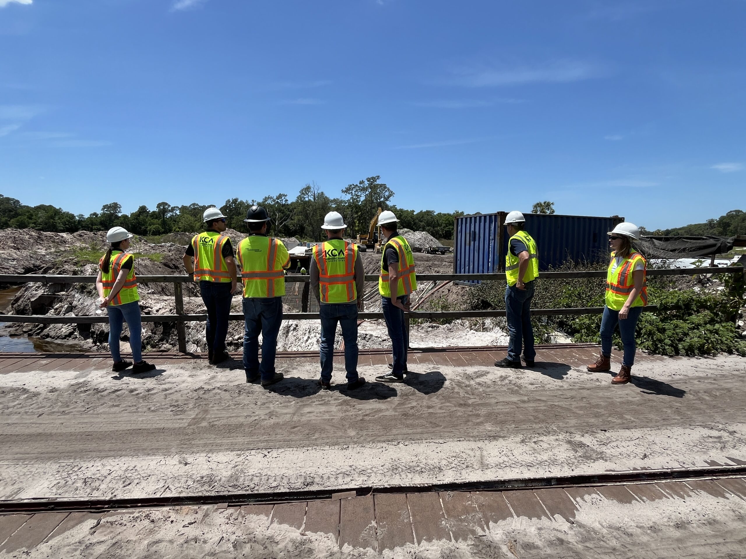 Seven people in safety gear stand on a sandy site, observing construction work