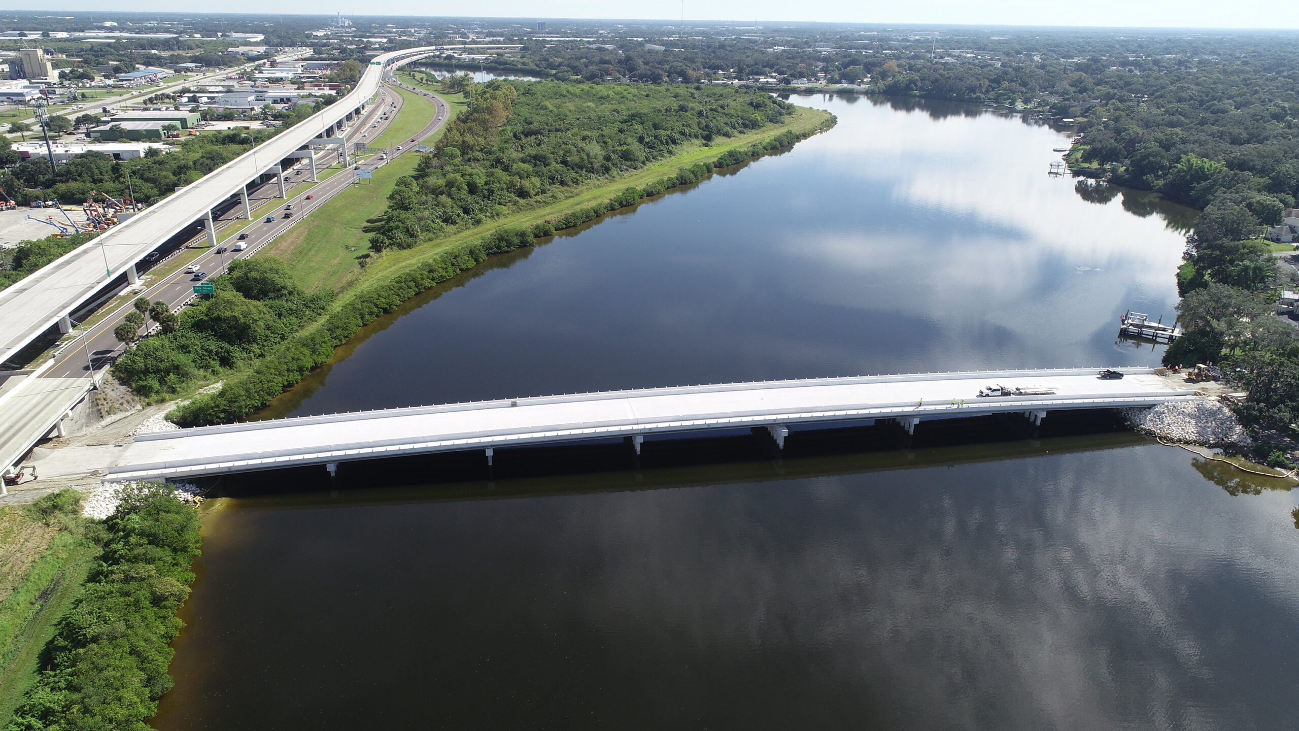 Aerial view of a newly constructed bridge spanning a wide river, surrounded by greenery and roads on a sunny day