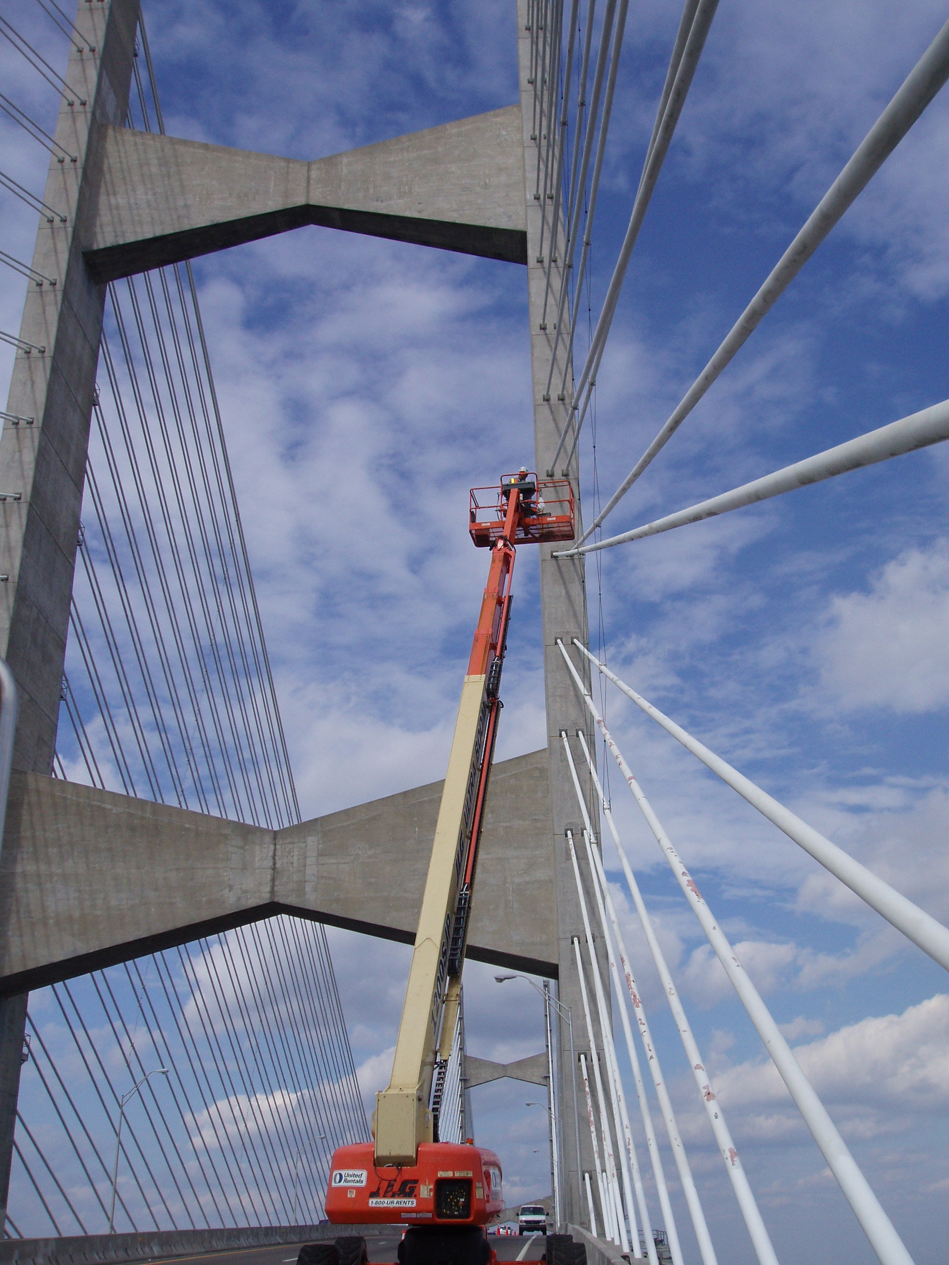 Person elevated on a lift repairing cables of a large suspension bridge under a blue sky