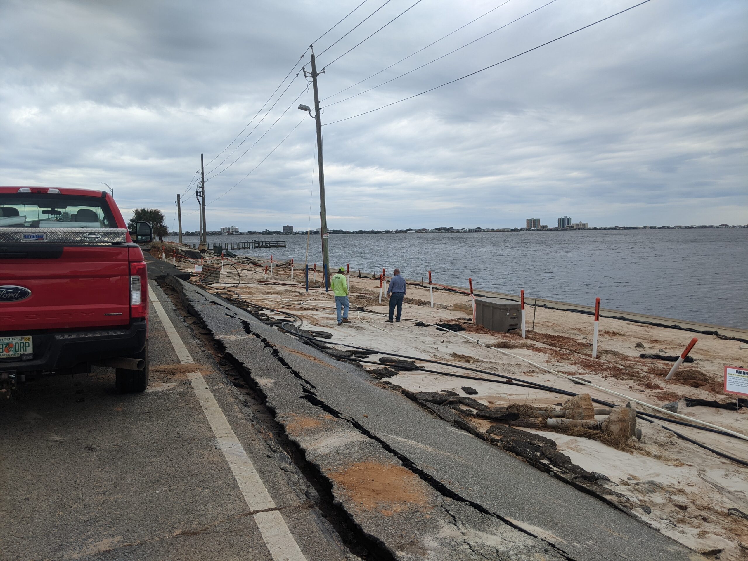 Damaged waterfront road with large cracks, red truck, two workers assessing site