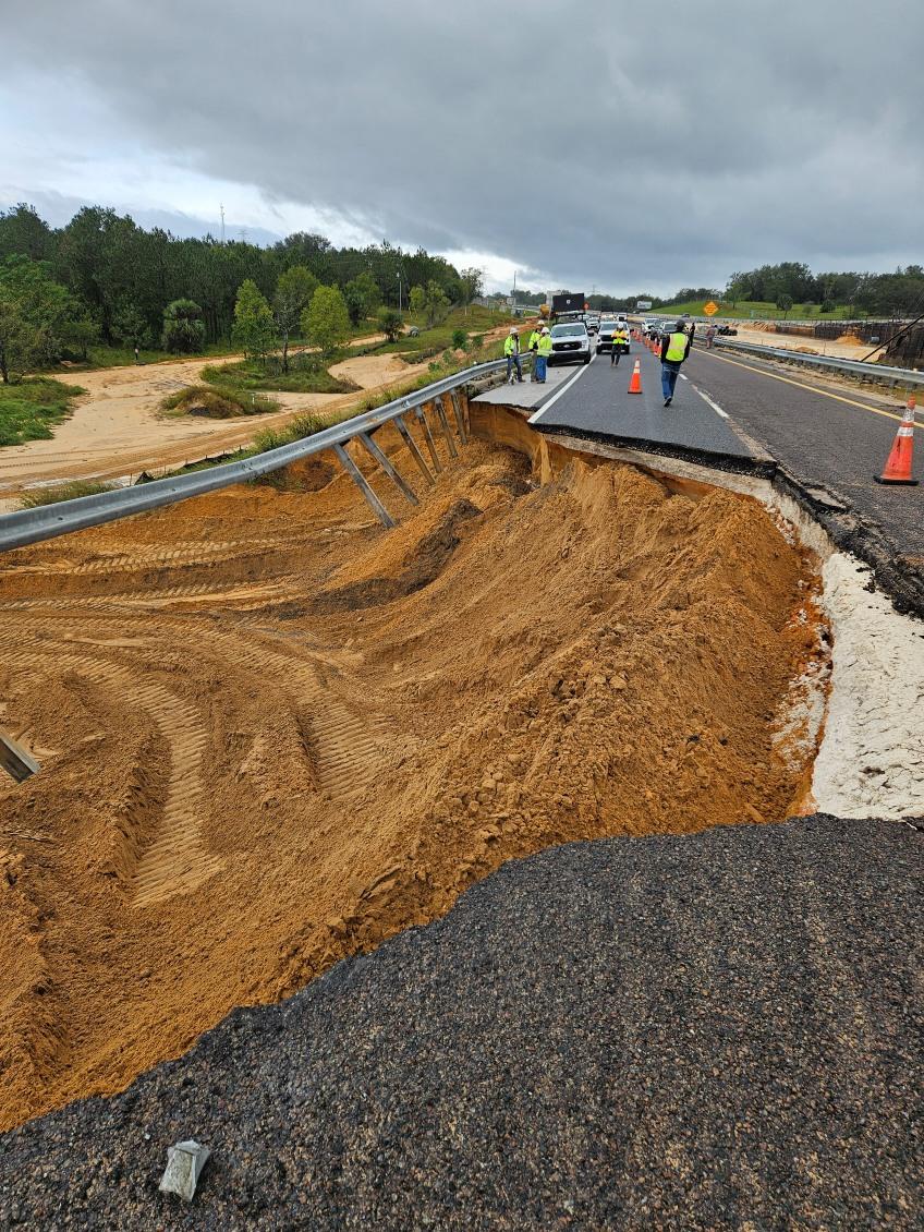 Severe road washout with exposed dirt; workers and vehicles on partially closed highway