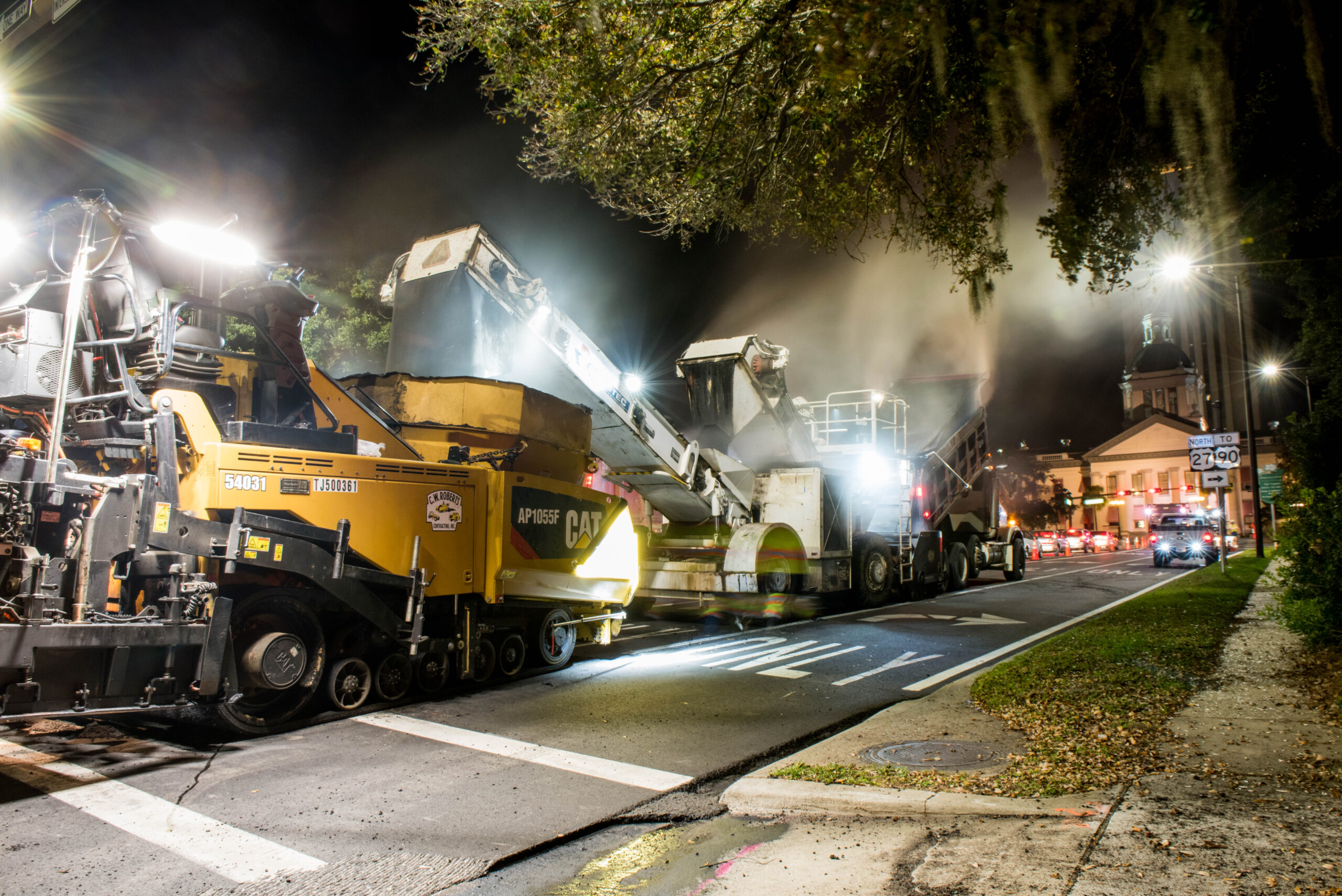 Nighttime road construction with heavy machinery, including yellow CAT AP1055F asphalt paver, active under bright lights