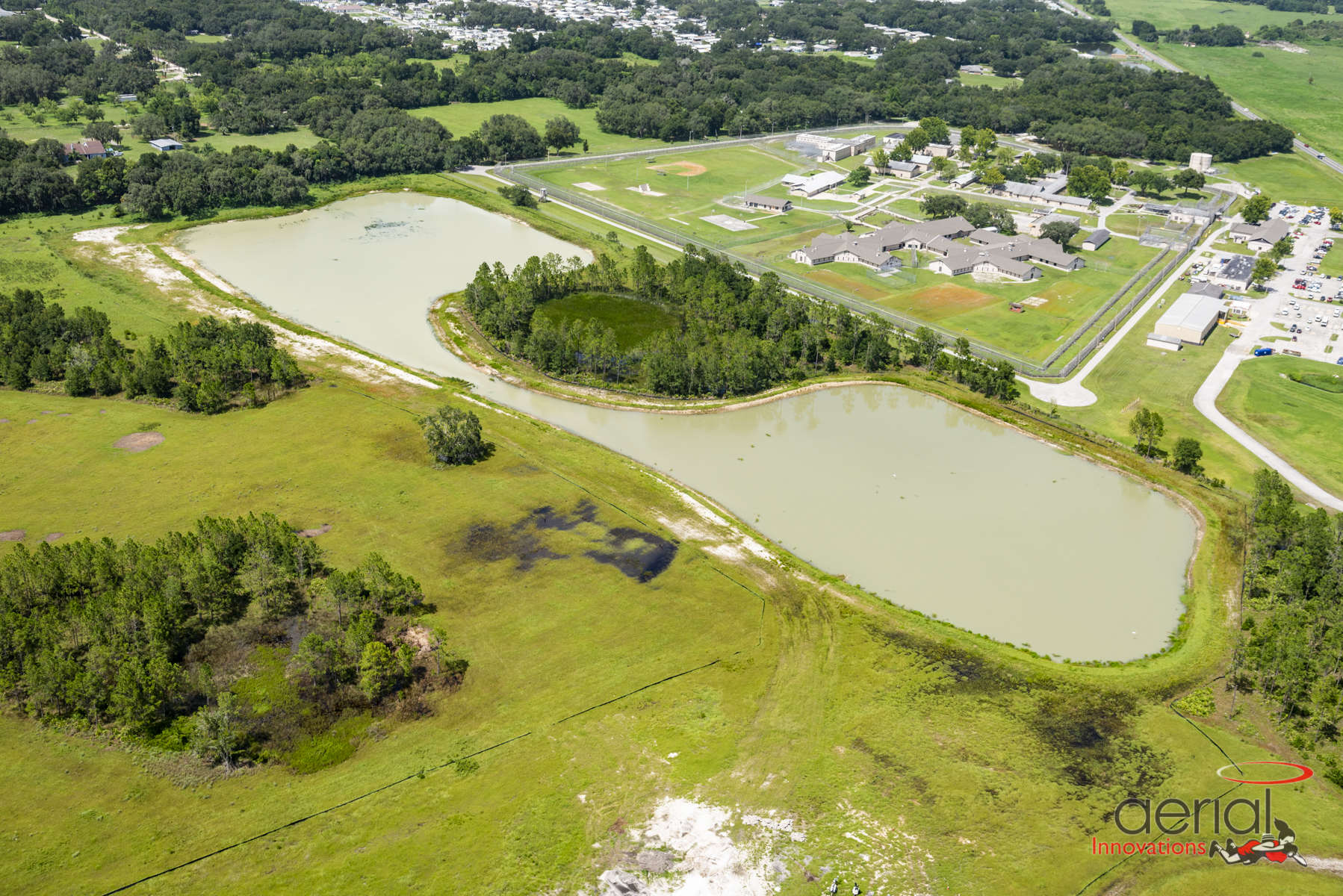 Aerial view of grassy landscape with two large ponds, surrounded by forested areas and buildings in the background