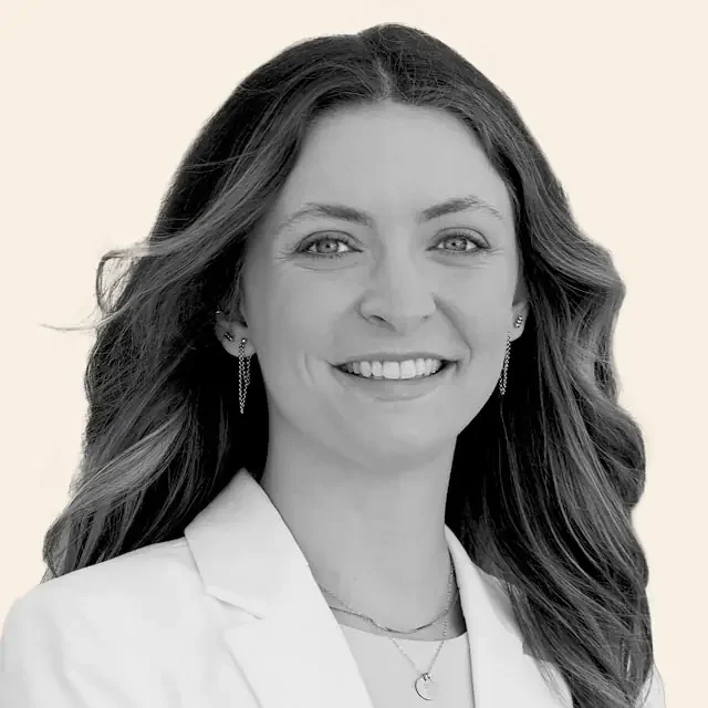 Ainsley smiling, wearing a white blazer, earrings, and necklaces against an off-white background.