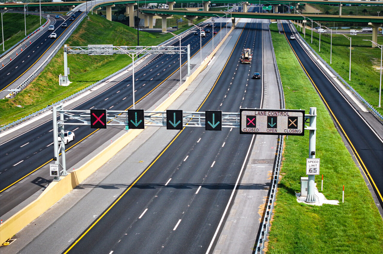 Highway with lane control signs; green arrows and red X indicating lane status. Speed limit 65 mph