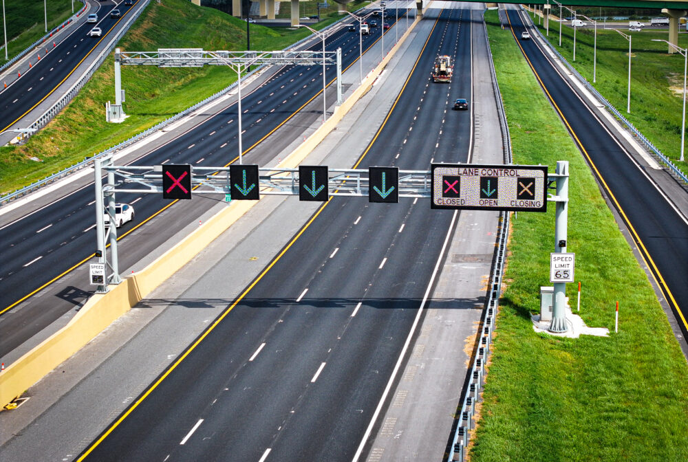 Highway with lane control signs; green arrows and red X indicating lane status. Speed limit 65 mph