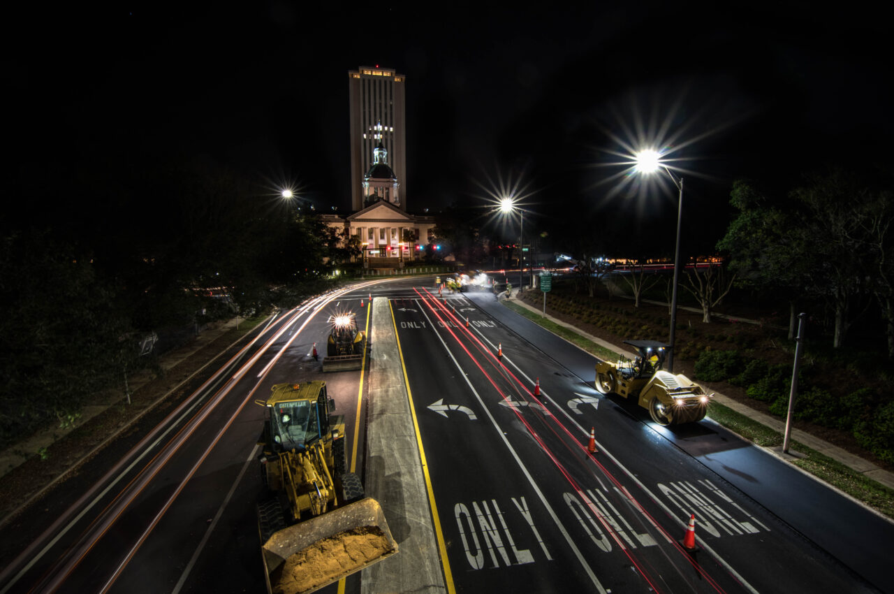 Night road construction in Tallahassee, Capitol building illuminated, vehicles with headlights, lane markings