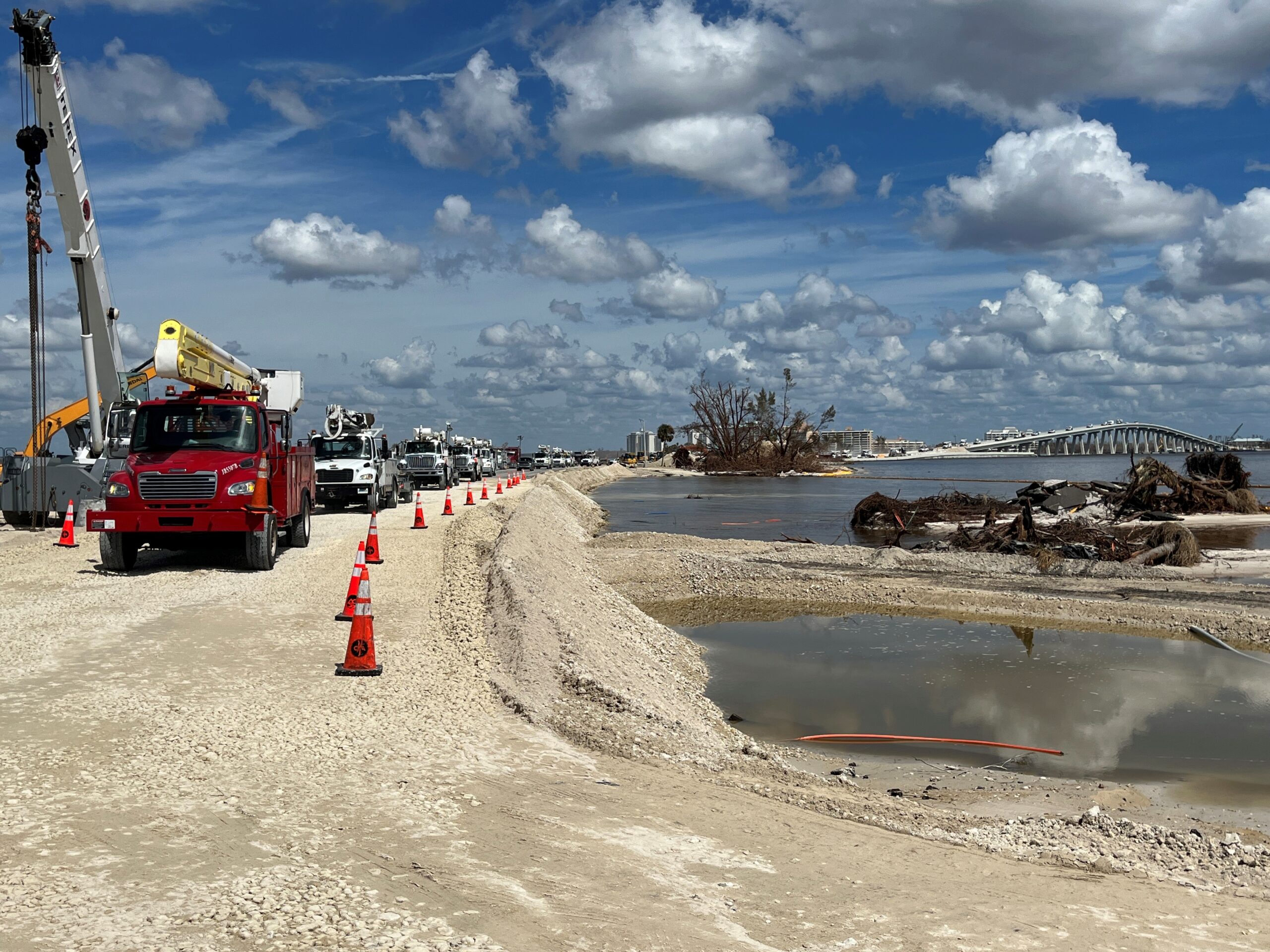 Emergency road repair on Sanibel Causeway with utility trucks and scattered debris under a partly cloudy sky