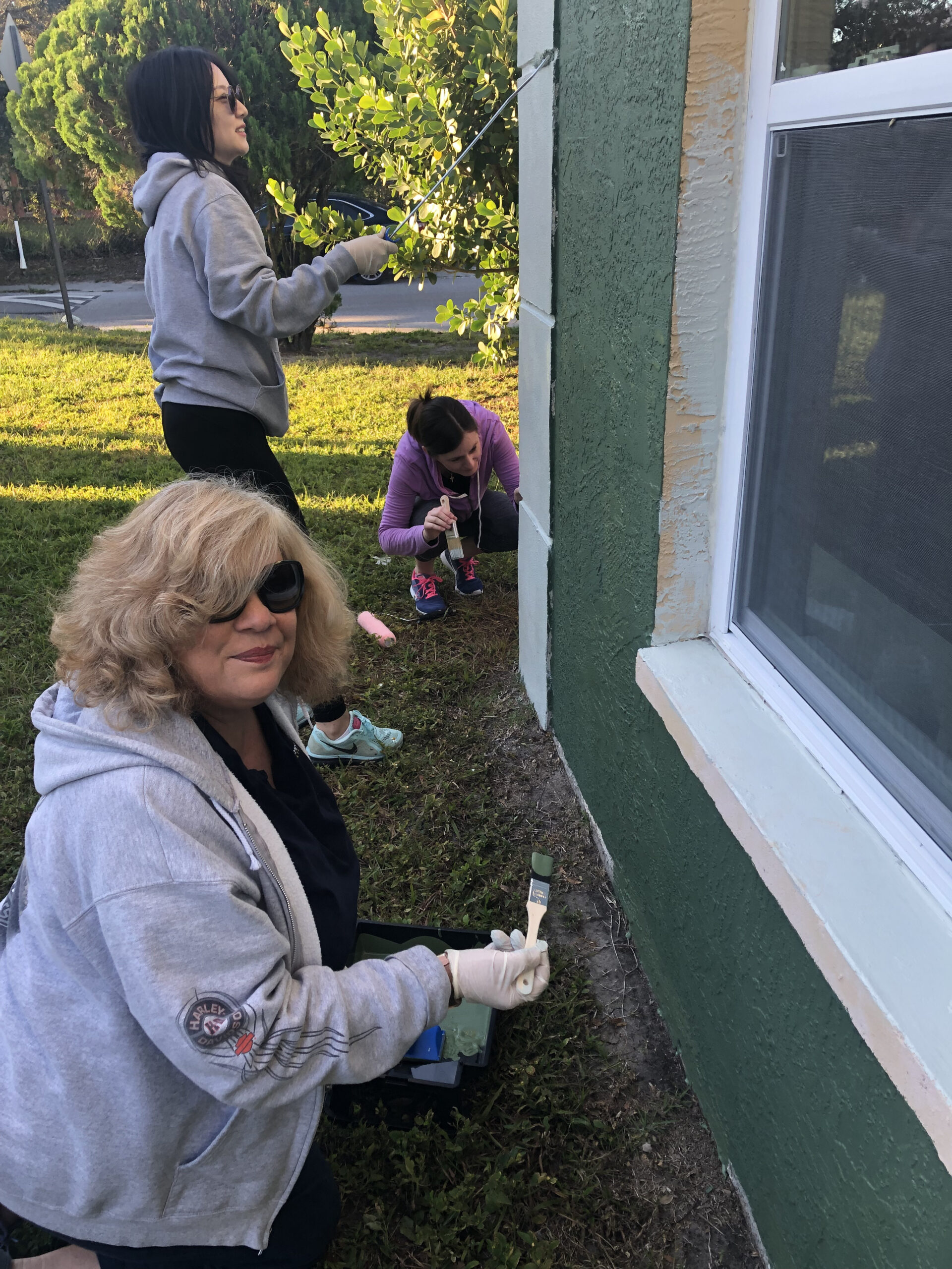 Three people painting an exterior wall green, wearing casual clothes and gloves