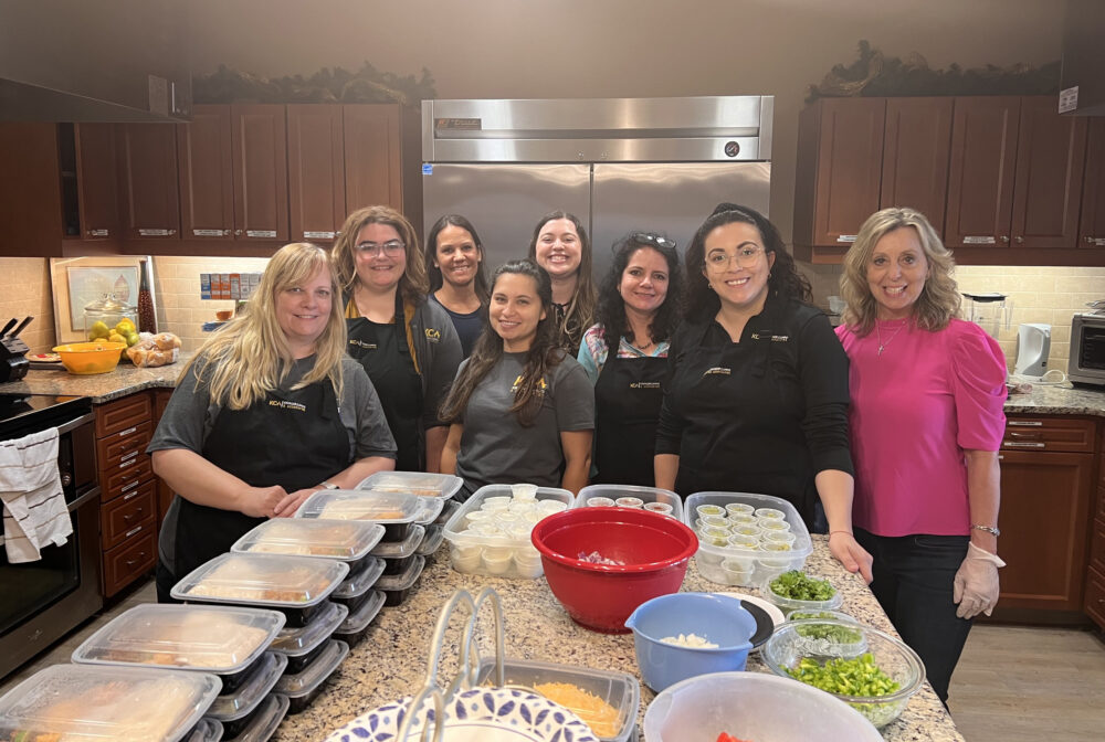Group of eight women in a kitchen preparing meals, smiling at the camera