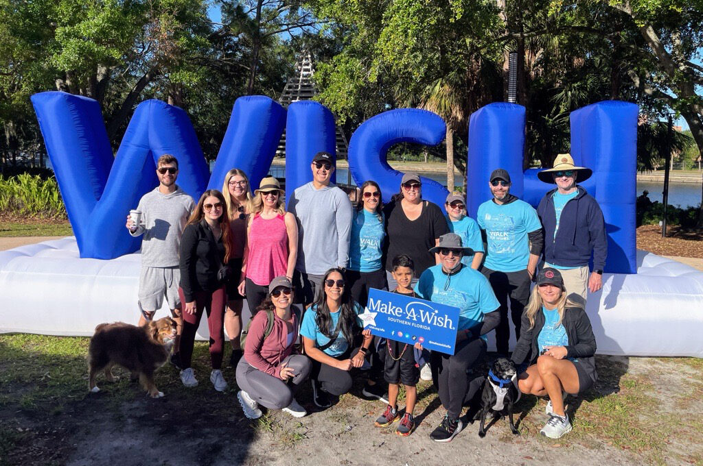 Group at "Walk for Wishes" event holding Make-A-Wish Southern Florida sign, smiling in front of large inflatable "WISH" letters