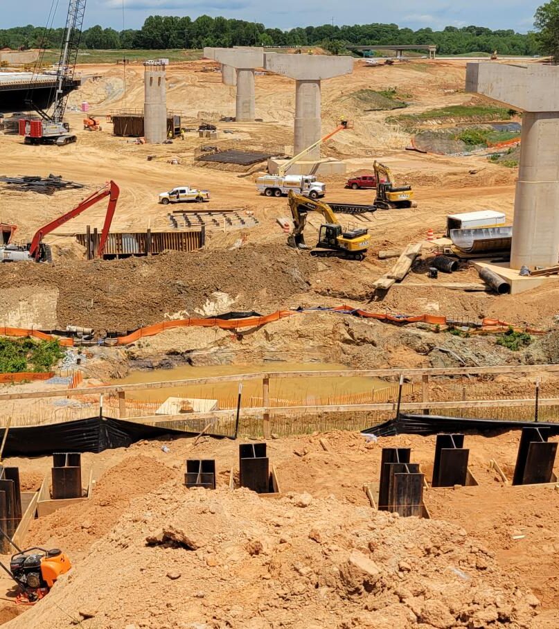 Construction site with workers and machinery, large concrete bridge columns, and excavation activity