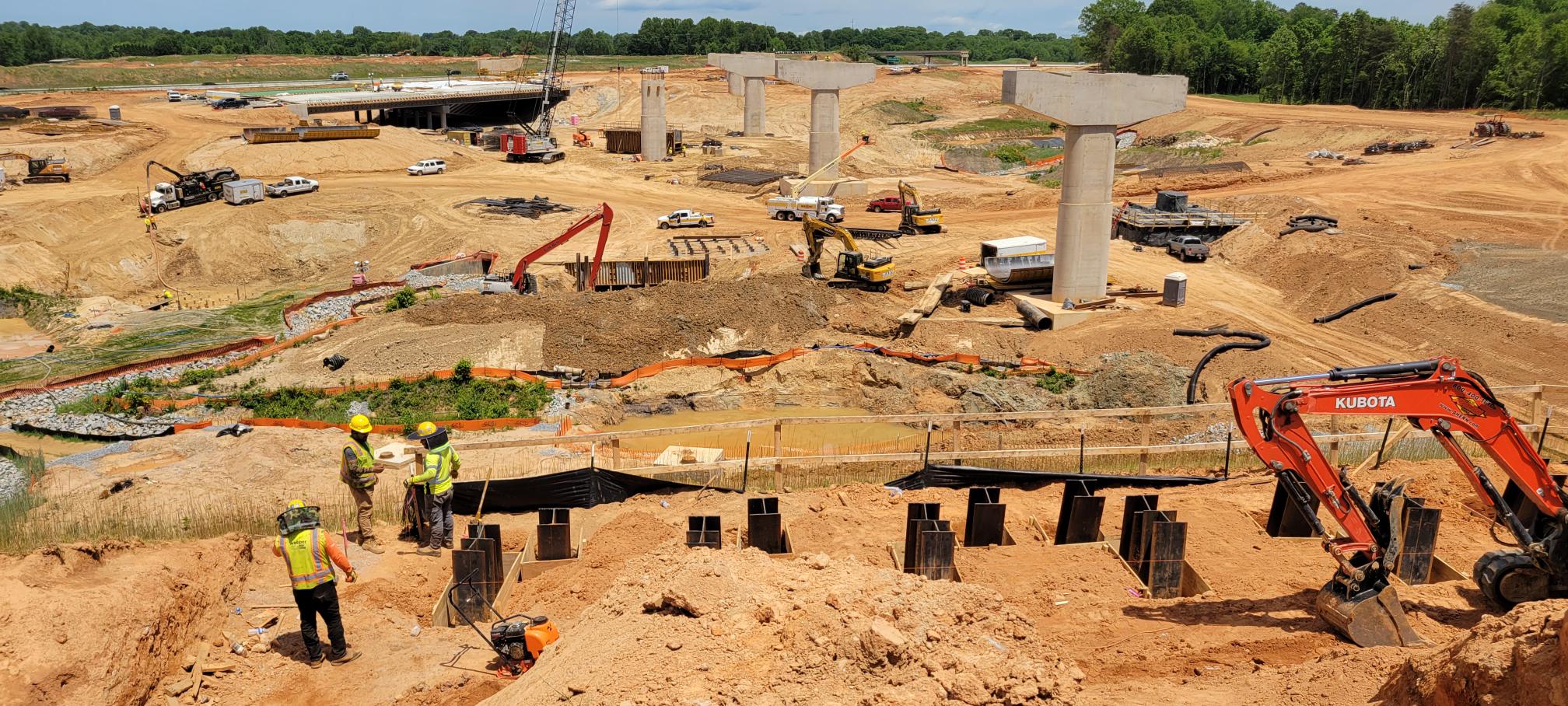 Construction site with workers and machinery, large concrete bridge columns, and excavation activity
