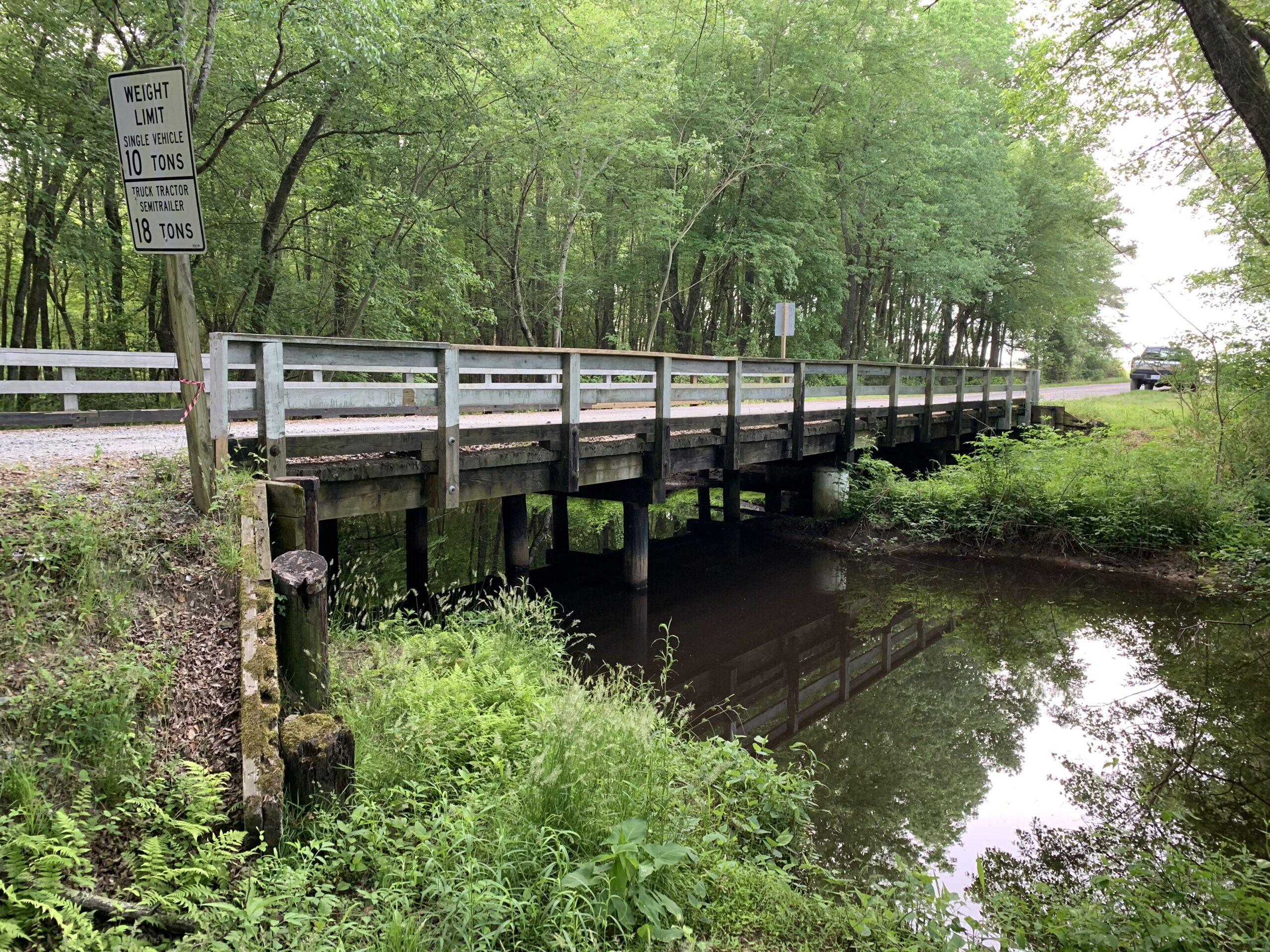Wooden bridge over creek surrounded by lush greenery, with a sign displaying weight limits for vehicles