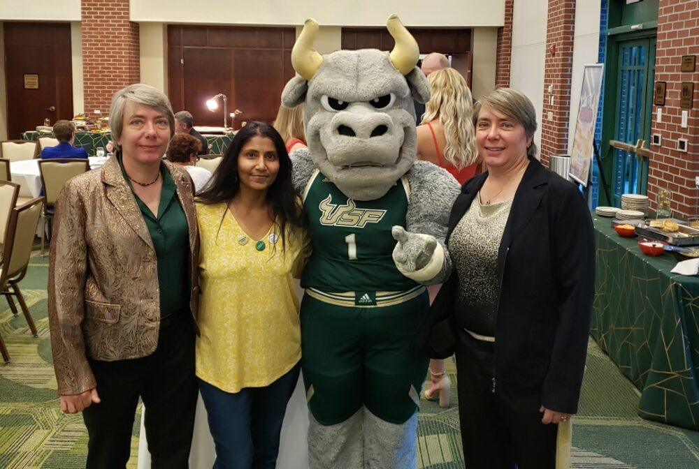 A mascot in USF uniform poses with three women, smiling in a conference room