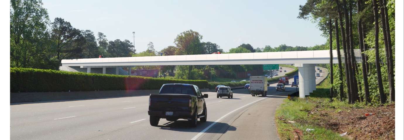 Highway under a white pedestrian bridge surrounded by trees, with light traffic on road