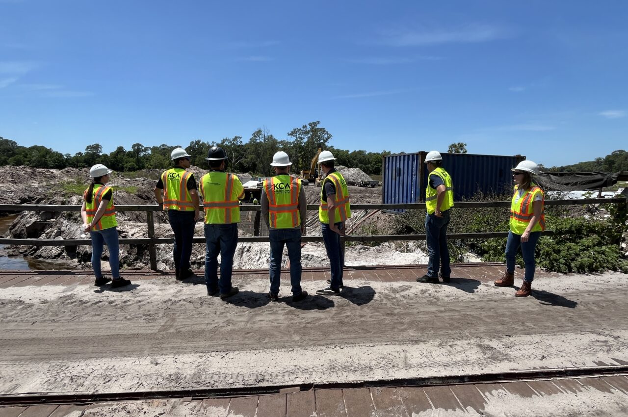 Seven construction workers in yellow safety vests and helmets discuss a site plan outdoors