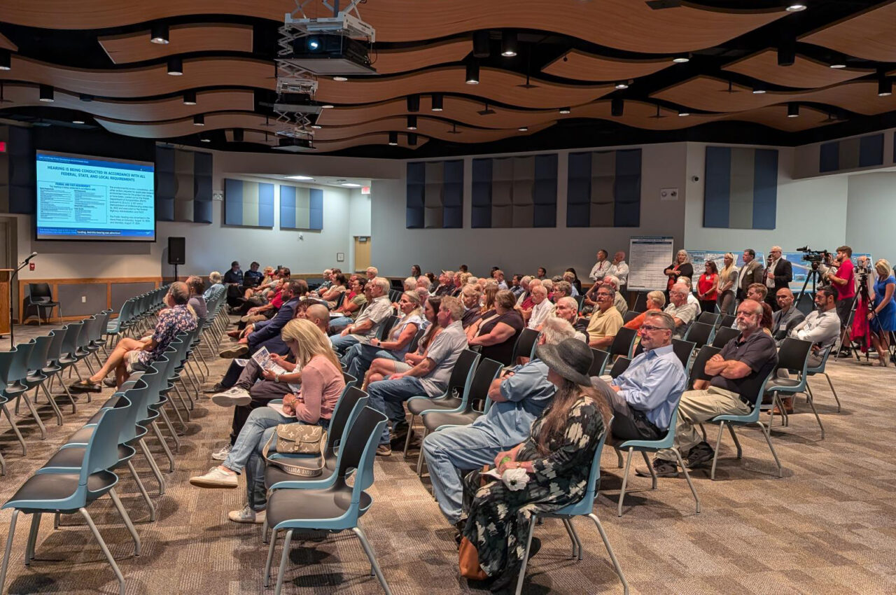 Audience seated at a public meeting, engaged with a speaker's presentation on a screen