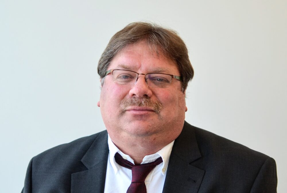 Darren wearing a suit and tie, standing against a plain background