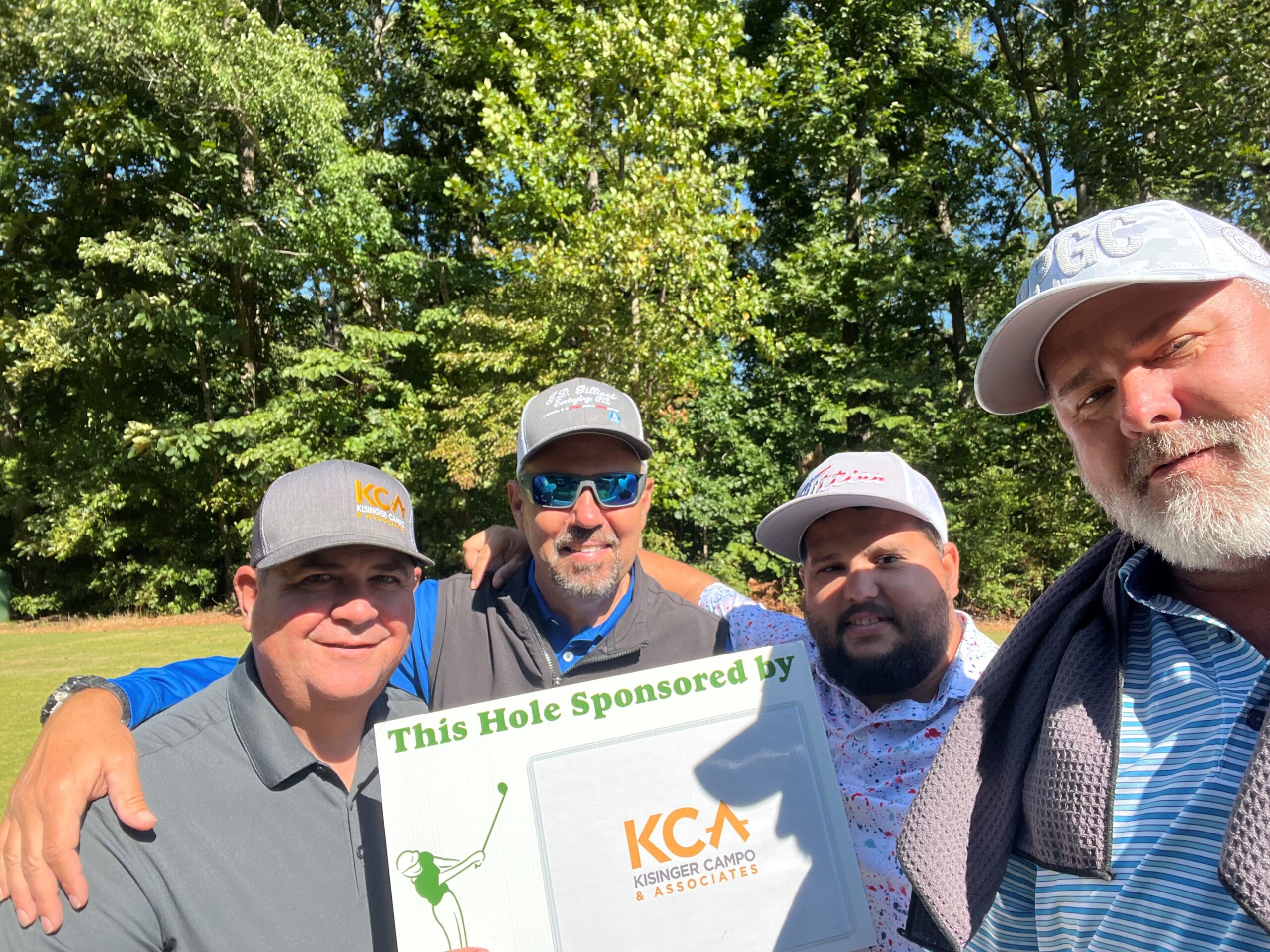 Four men standing with a sign reading "This Hole Sponsored by KCA" on a golf course