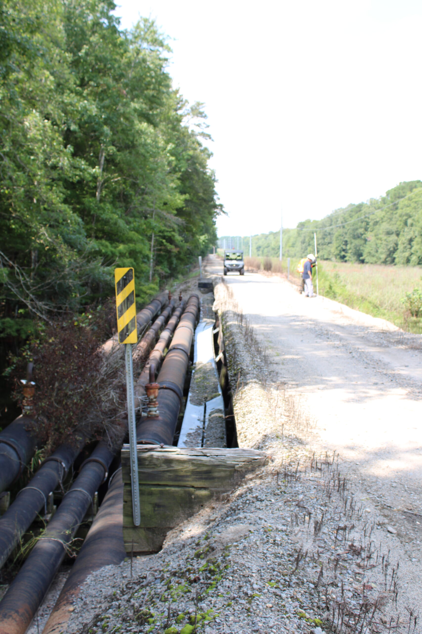 Gravel road with large pipelines; worker in helmet measuring, vehicle approaching in distance