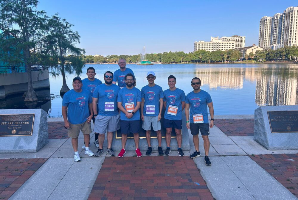 Group of eight men in matching blue shirts posing by a lakeside in Orlando, smiling, wearing race bibs, with cityscape background