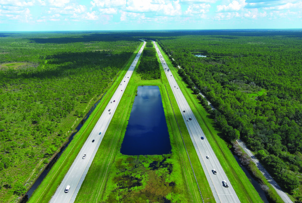 Aerial view of a divided highway with a water canal and dense green forest surrounding it