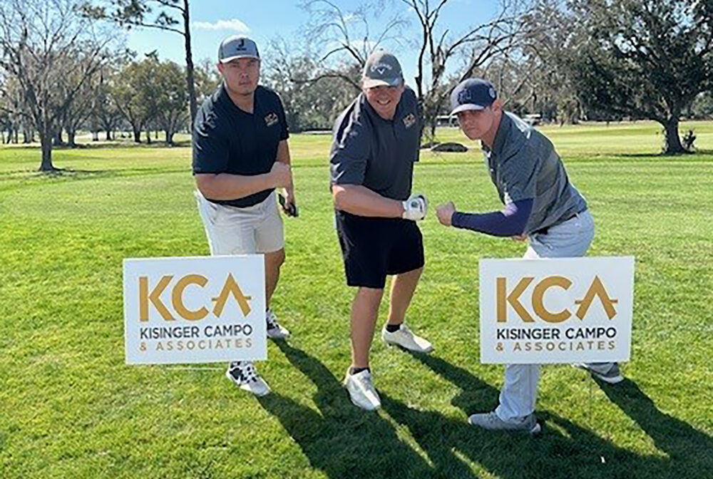 Three men pose together on a golf course, surrounded by KCA Kisinger Campo & Associates signs
