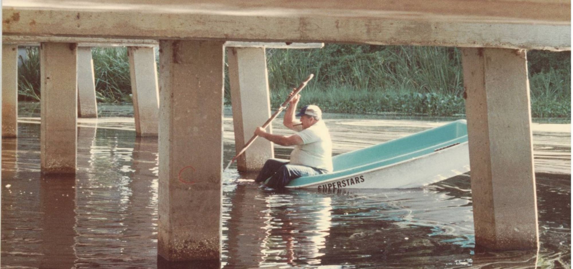 Ed paddles a small boat labeled "SUPERSTARS" under a bridge in calm water