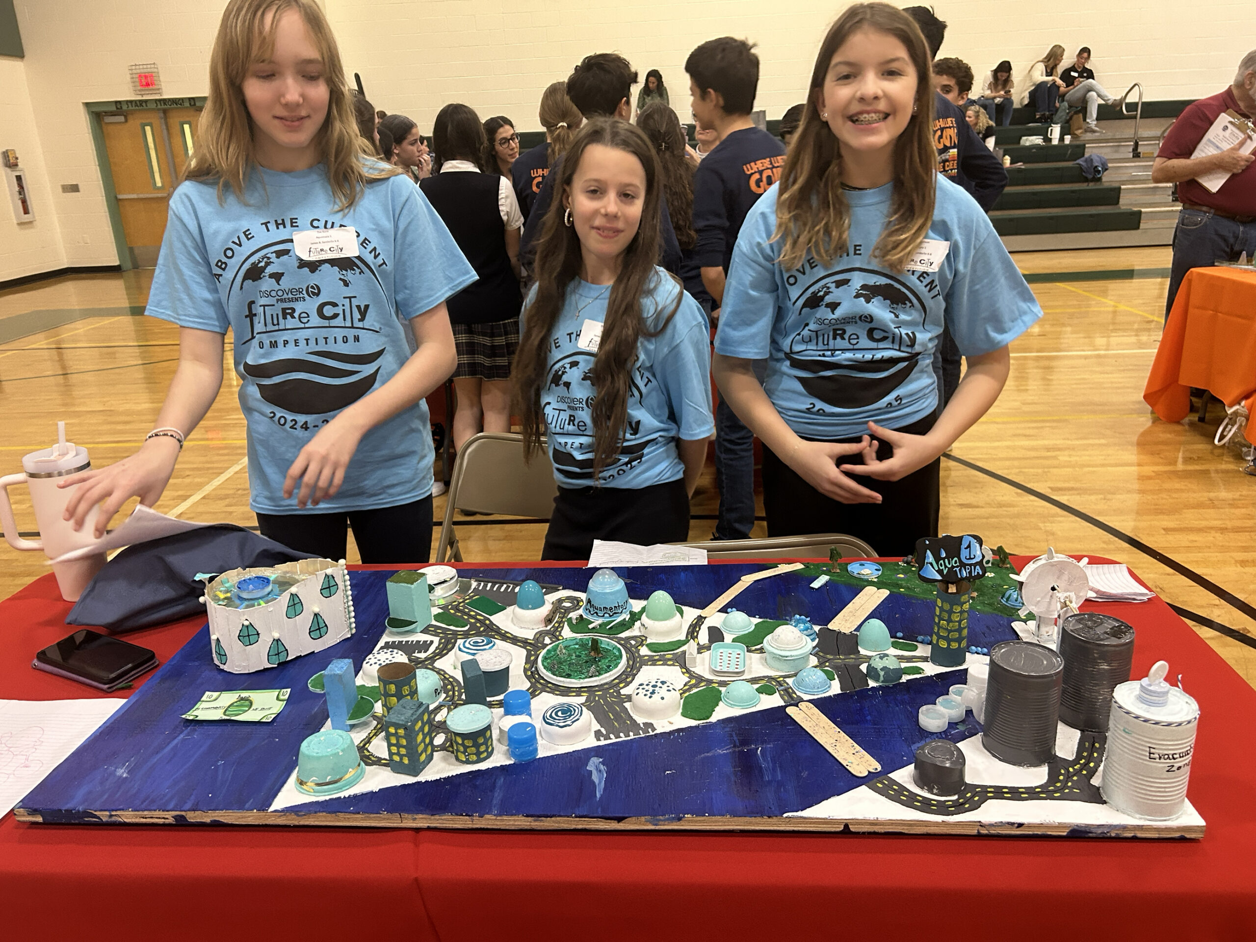 Three girls in blue shirts presenting a model city at a competition table