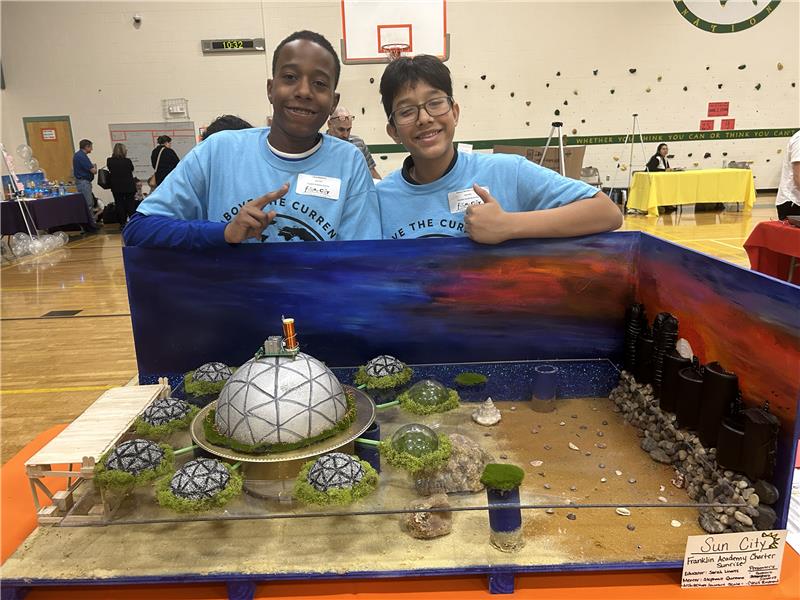 Two students showcase a futuristic city model with domed structures, wearing blue shirts in a gymnasium setting