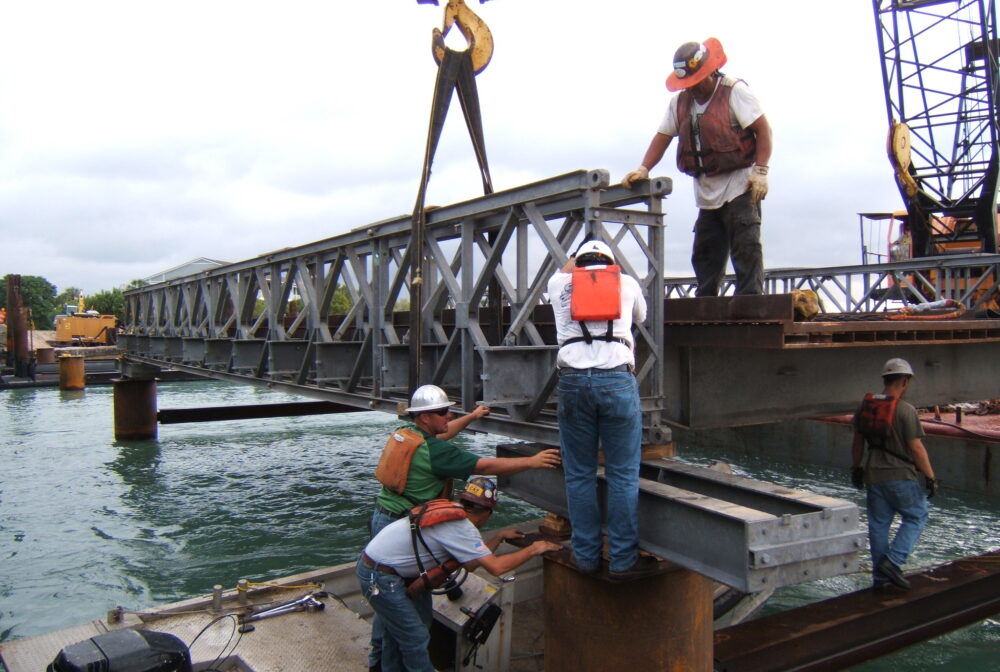Workers in safety gear positioning bridge section over water using a crane