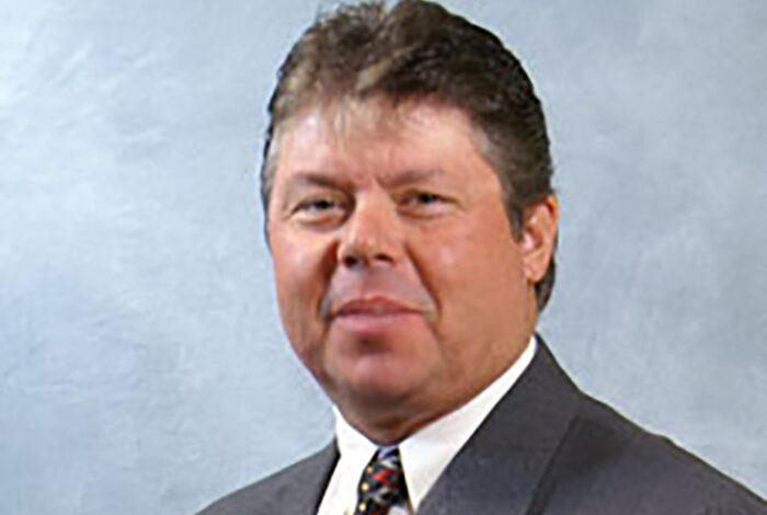 Gott Ron wearing a suit and tie against a light gray background