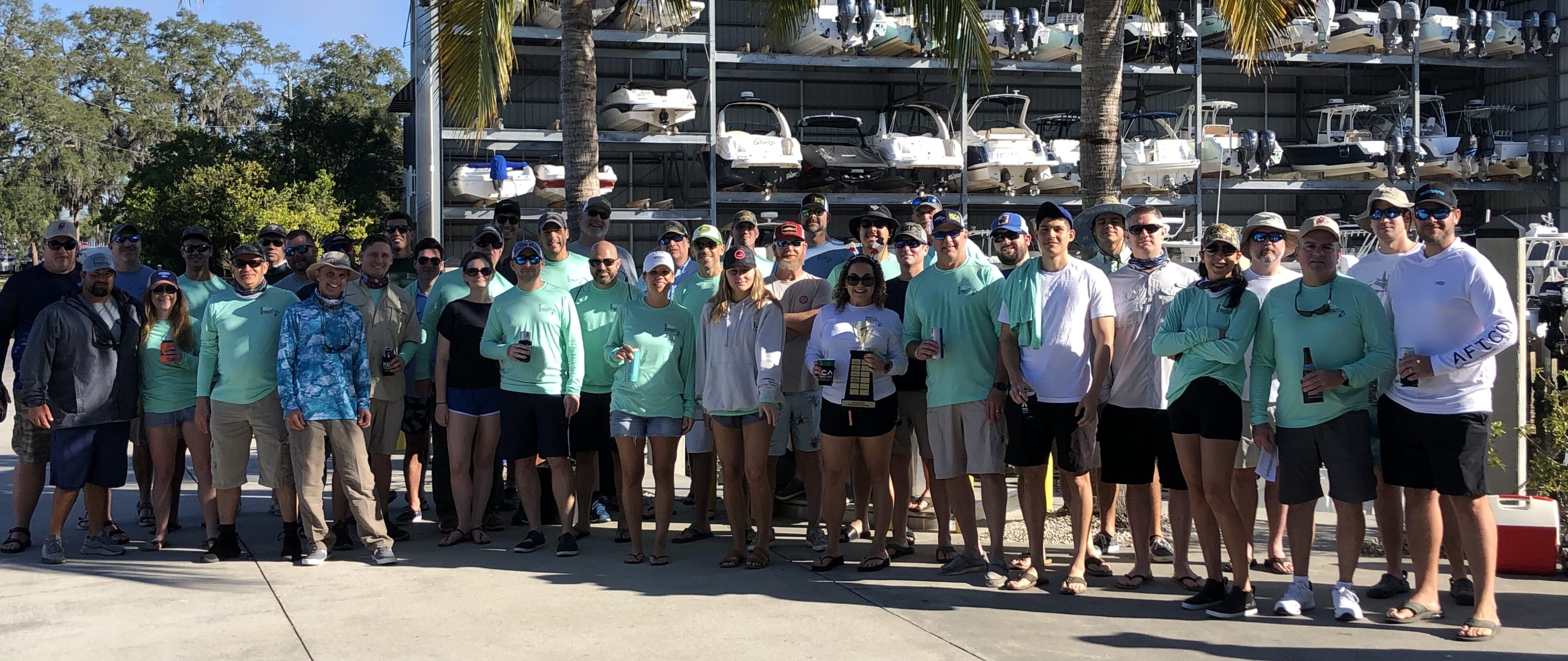 Group of people standing outdoors in front of boats, with palm trees and wearing casual tropical clothing