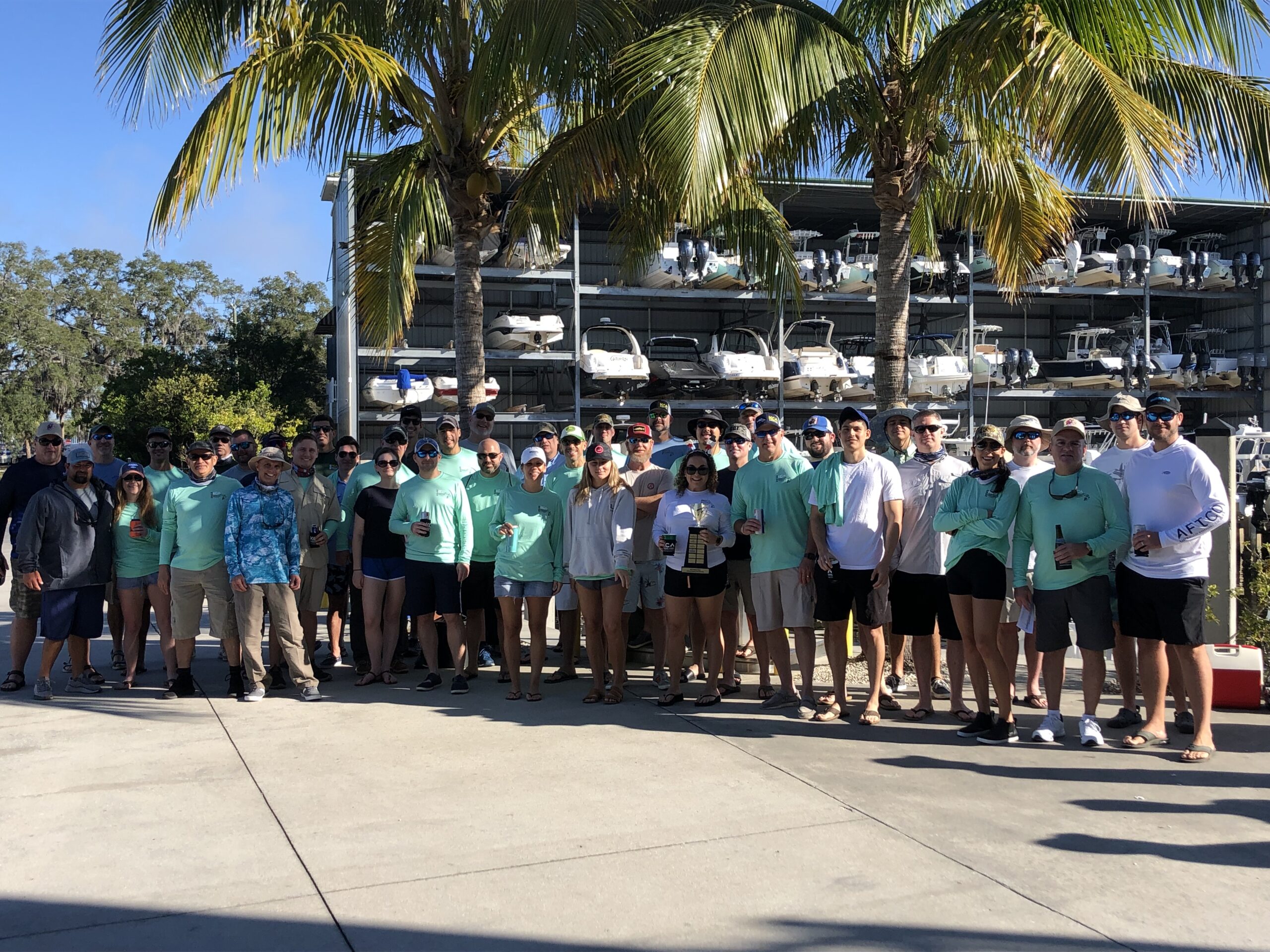Group of people standing outdoors in front of boats, with palm trees and wearing casual tropical clothing