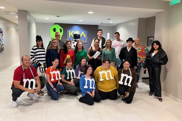 Group of people in various Halloween costumes posing in an office lobby, some dressed as M&M candies