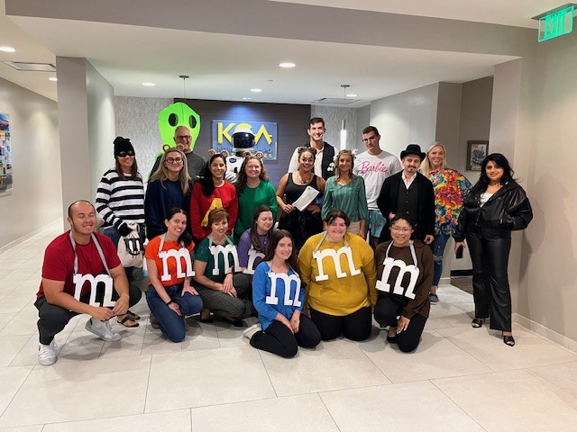 Group dressed in various costumes, including M&M characters, standing in an office setting, smiling and posing together