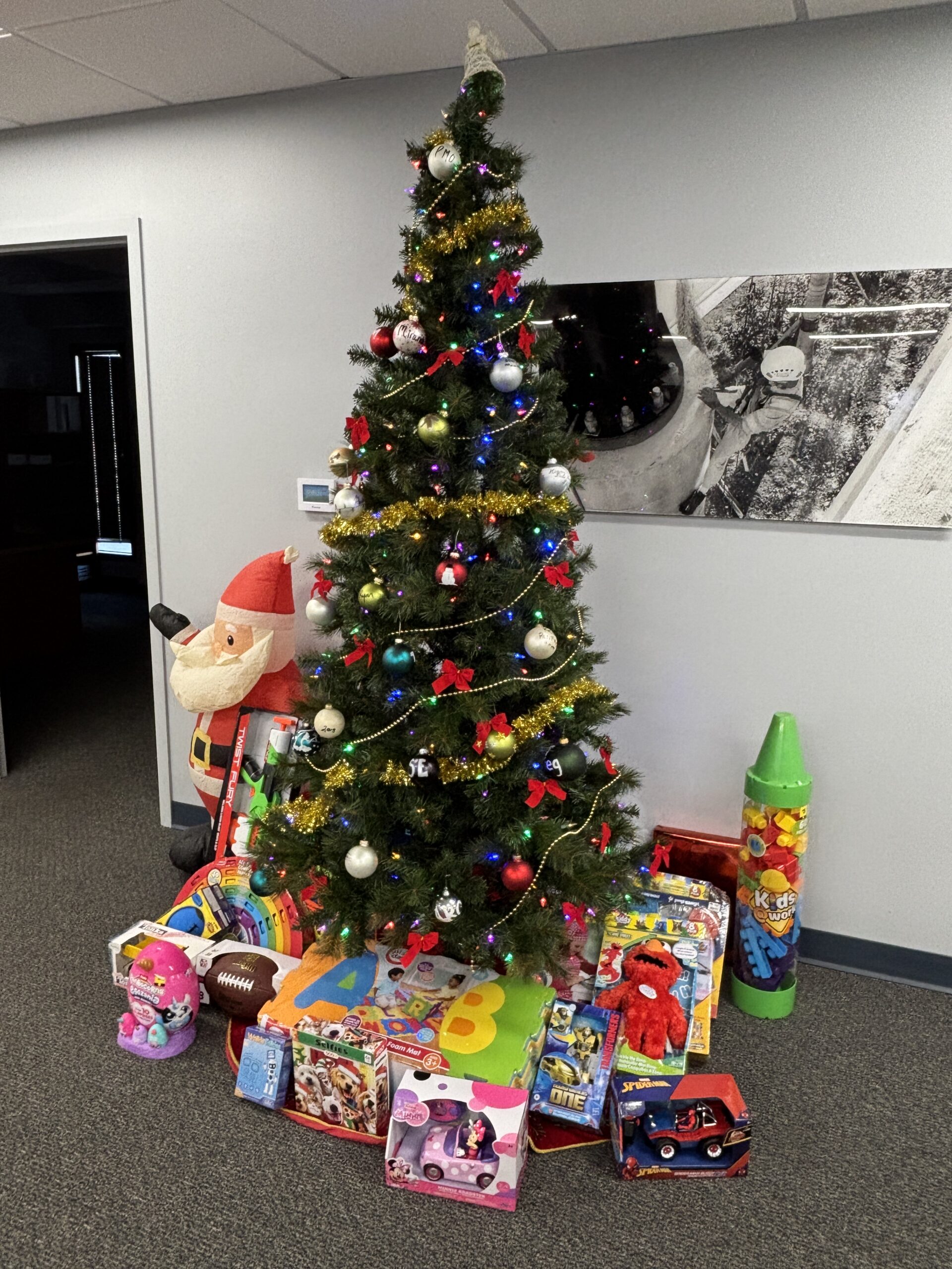 Christmas tree with lights and ornaments, surrounded by wrapped gifts and toys, including a Santa decoration and a large crayon