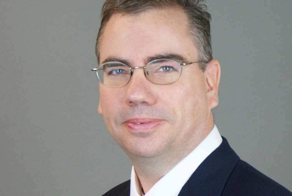 Hubert David smiling in a suit and tie against a gray background
