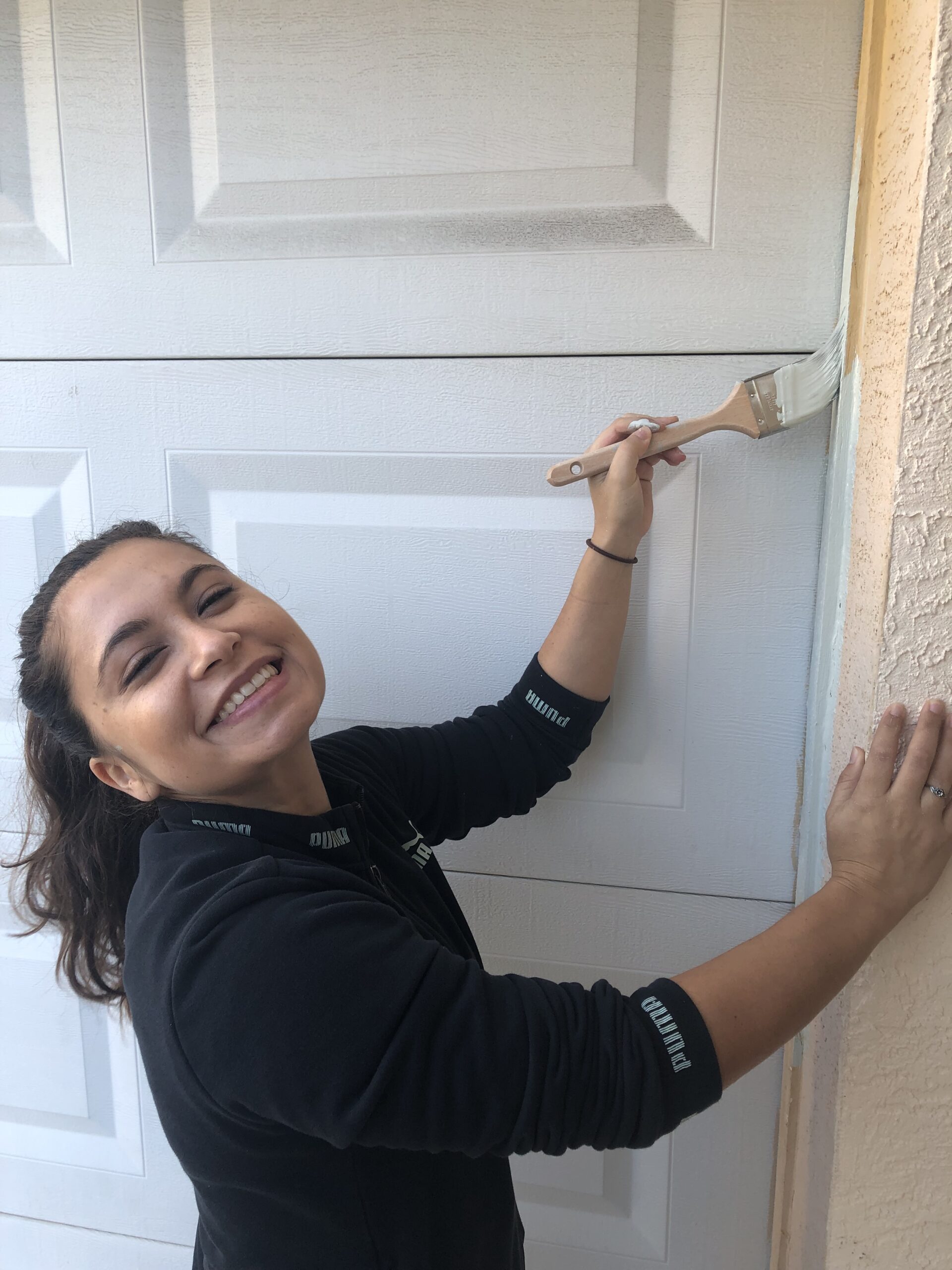Smiling woman painting a garage door edge with a brush, wearing a black Puma jacket