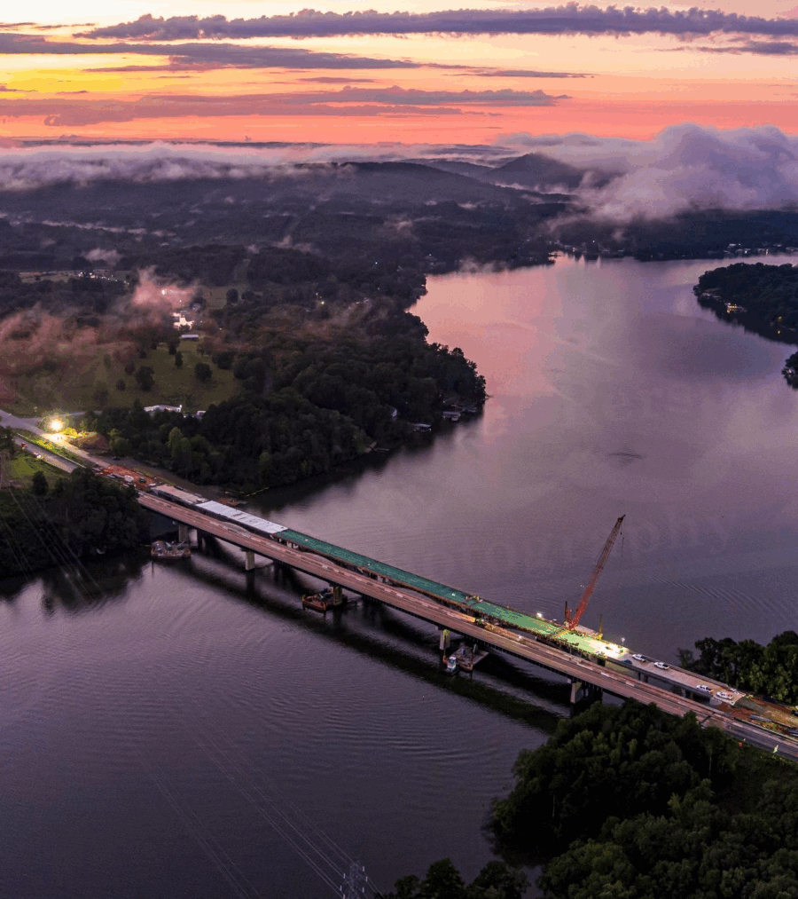 Aerial view of a bridge construction site over a river at sunset, surrounded by fog and lush trees